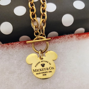 Pode incluir: Colar de ouro com um pingente de Mickey Mouse que diz "Por favor, devolva para Mickey & Co. Main Street, USA 1928".