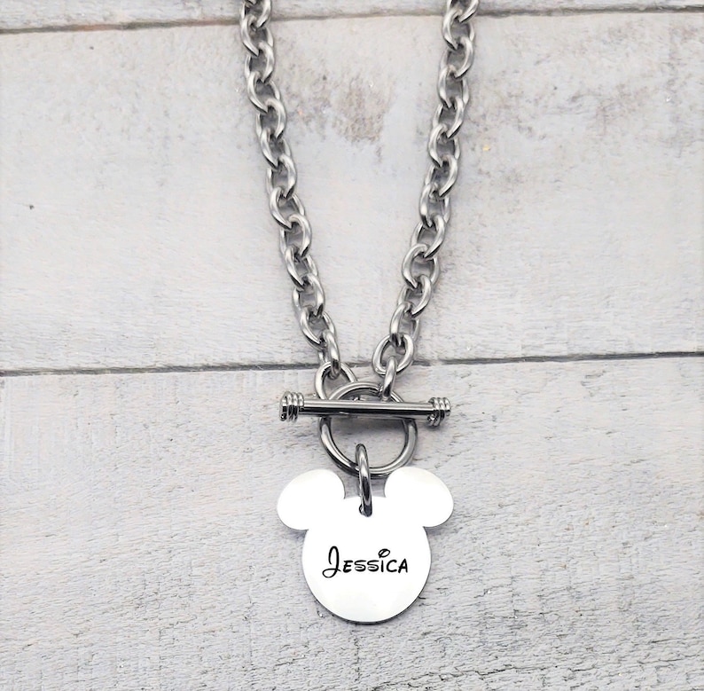 Pode incluir: Um colar de prata com fecho de correr e um pingente em forma de Mickey Mouse com o nome "Jessica" gravado.