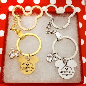 May include: Two gold and silver Mickey Mouse keychains with charms. Each keychain features a Mickey Mouse head charm with the text "Please return to Mickey & Co. Main Street, USA 1928".