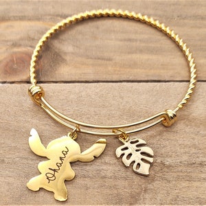 Puede incluir: Pulsera de brazalete de tono dorado con un dise&ntilde;o trenzado. La pulsera tiene dos abalorios: un personaje de Stitch con el nombre "Shana" grabado en &eacute;l y un abalorio de hoja tropical de tono dorado.