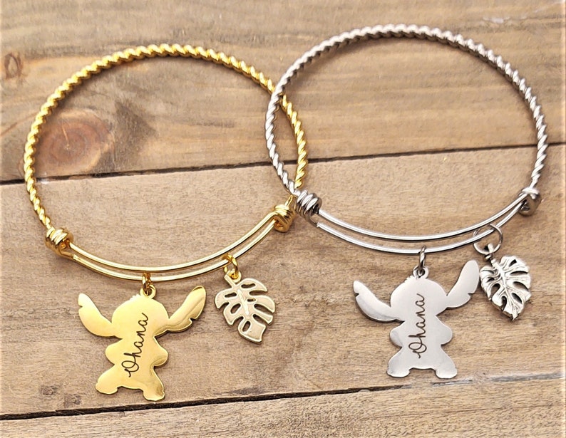 Puede incluir: Dos pulseras de brazalete de oro y plata con dijes. Los dijes son un personaje de dibujos animados con la palabra "Ohana" grabada en &eacute;l y una hoja tropical.