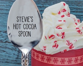 May include: A silver spoon with the text "Stevie's Hot Cocoa Spoon" engraved on the front. The spoon is next to a red mug with white snowflakes around the rim, filled with whipped cream and red and white candy sprinkles.
