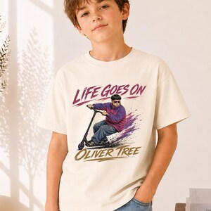 May include: A cream-colored short-sleeve t-shirt featuring a graphic print of a person riding a scooter. The text "LIFE GOES ON" appears in red above the figure, and "OLIVER TREE" is printed in gold below, with purple paint-like accents.