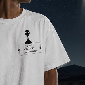 May include: White t-shirt with a black alien graphic holding a sign that reads "I DON'T BELIEVE IN HUMANS." The shirt is a casual style, perfect for everyday wear. The background features a night sky with a UFO.