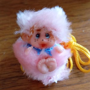May include: A small pink plush doll with a yellow cord. The doll is wearing a pink hat and a blue and white outfit. The doll has a painted face with freckles.