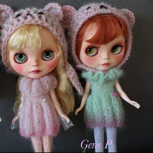 Può includere: Due bambole con grandi occhi e guance rosate indossano cuffiette lavorate a maglia rosa e viola chiaro con orecchie da orso. La bambola a sinistra ha capelli biondi e un vestito rosa, quella a destra capelli ramati e un vestito verde menta. Testo Gene P.