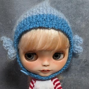 May include: A doll with blonde hair and freckles wearing a light blue and medium blue crocheted bonnet with small side ear details and a single blue button closure under the chin, paired with a white shirt featuring red striped long sleeves.