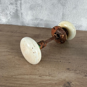 Może przedstawiać: Antyczny zestaw gałek do drzwi z dwoma owalnymi, kremowymi porcelanowymi gałkami. Metalowe elementy wykazują oznaki rdzy i zużycia, wskazujące na wiek. Zestaw zawiera trzpień i elementy montażowe.