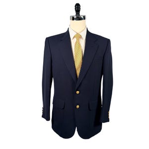 Puede incluir: Un blazer azul marino con botones dorados y una corbata amarilla estampada. El blazer tiene dos bolsillos delanteros y un bolsillo en el pecho. La chaqueta se exhibe en un maniquí.