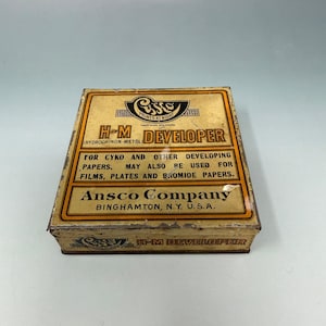 Peut inclure: Boîte vintage de révélateur Ansco H-M. La boîte rectangulaire a un fond beige avec du texte noir et orange. Le texte comprend "H-M DEVELOPER" et "Ansco Company, Binghamton, N.Y. U.S.A.". La boîte est pour développer des films, des plaques et des papiers bromure.