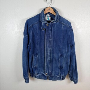 May include: A medium-wash blue denim jacket featuring a fold-down collar, a front zipper closure, and a buttoned throat latch. The jacket includes two vertical front pockets, long sleeves with buttoned cuffs, and a ribbed hem. A Trader Bay label is visible.