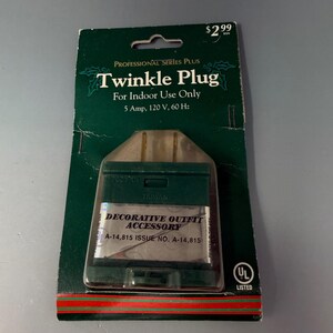 May include: A green and clear plastic Twinkle Plug for indoor use, packaged on a dark green card. The packaging includes the text "Twinkle Plug" and "Decorative Outfit Accessory". The plug is rated at 5 Amp, 120 V, 60 Hz.