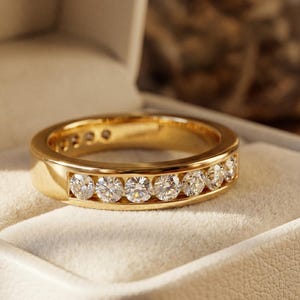 Peut inclure: Une bague en or avec un sertissage en canal, ornée d'une rangée de diamants étincelants. La bague est présentée dans un écrin blanc, sur un fond doux et neutre. Le design de la bague est simple mais élégant.