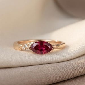 Könnte beinhalten: Ein Ring aus Roségold mit einem zentralen, tiefroten Edelstein im Marquise-Schliff. Die Ringschiene ist mit zwei kleinen, klaren, runden Akzentsteinen auf einer Seite der Fassung versehen, was dem Schmuckstück ein feines Aussehen verleiht.