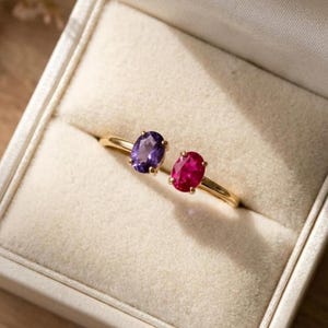 Puede incluir: Un anillo abierto de oro con dos piedras preciosas de corte ovalado, una amatista morada y un rubí rosa vibrante, engastadas en una banda de metal fina. El anillo se muestra sobre un cojín de terciopelo color crema en un joyero.