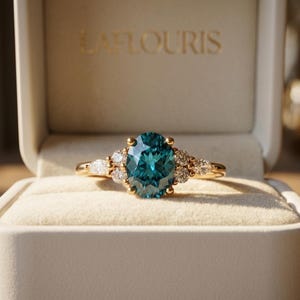 Puede incluir: Un anillo de oro con una gran piedra preciosa ovalada de color azul verdoso en el centro, flanqueada por diamantes blancos más pequeños de talla redonda y pera a cada lado. El anillo se presenta en una caja beige con el nombre LAFLOURIS en letras doradas.
