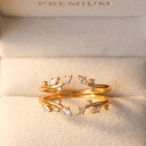 Peut inclure: Une bague ouverte en or composée de deux anneaux parallèles en forme de branches, ornés de pierres claires taille marquise et ronde. La bague est présentée dans un écrin de couleur claire avec le mot PREMIUM gravé sur le couvercle intérieur.
