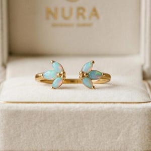 May include: A gold open ring featuring two symmetrical clusters of marquise-cut opals, each set with three stones arranged in a leaf-like pattern. The ring is displayed inside a cream-colored jewelry box with the word NURA printed in gold.