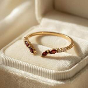 May include: A gold-colored open ring featuring three maroon-colored gemstones on each side, with small clear stones interspersed. The ring is presented in a white velvet jewelry box, showcasing its elegant design.