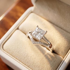 Könnte beinhalten: Ein silberner Verlobungsring mit einem großen, quadratisch geschliffenen Diamanten. Der Ring befindet sich in einer weißen Schachtel mit beigem Innenraum. Ein kleineres, gebogenes Band mit kleineren Diamanten liegt daneben.