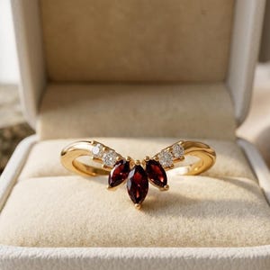 Peut inclure: Une bague en or en forme de V ornée de trois grenats rouge foncé taille marquise au centre, flanqués de deux petits diamants blancs ronds de chaque côté, le tout monté sur un anneau en or poli dans un écrin à bijoux beige.