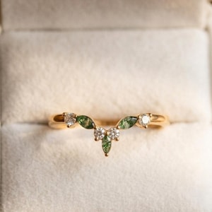 May include: A gold-colored ring with a curved design. The ring features small, round, clear gemstones and green, leaf-shaped stones. The ring is set against a soft, neutral background, highlighting its delicate details.