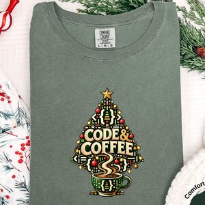 May include: A moss green t-shirt featuring a graphic of a Christmas tree formed by programming brackets and coffee-related elements. The text CODE & COFFEE is centered within the tree design, which sits above a green coffee mug.