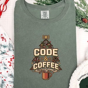 May include: A moss green crewneck t-shirt featuring a graphic of a Christmas tree shaped like a coffee cup with the text CODE & COFFEE. The shirt has a Comfort Colors label at the inner neckline and is displayed on a white surface.