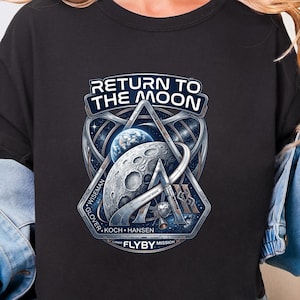 May include: Black crew neck t-shirt with a graphic that reads "RETURN TO THE MOON." The graphic features a detailed illustration of the moon, Earth, and a spacecraft, with the names of the crew members. The shirt is made of a soft material.