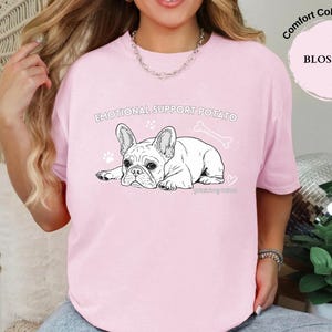 May include: A pale pink t-shirt featuring a black and white French Bulldog illustration with the text "EMOTIONAL SUPPORT POTATO". The shirt also has paw prints, a bone, and the word "BLOSSOM".