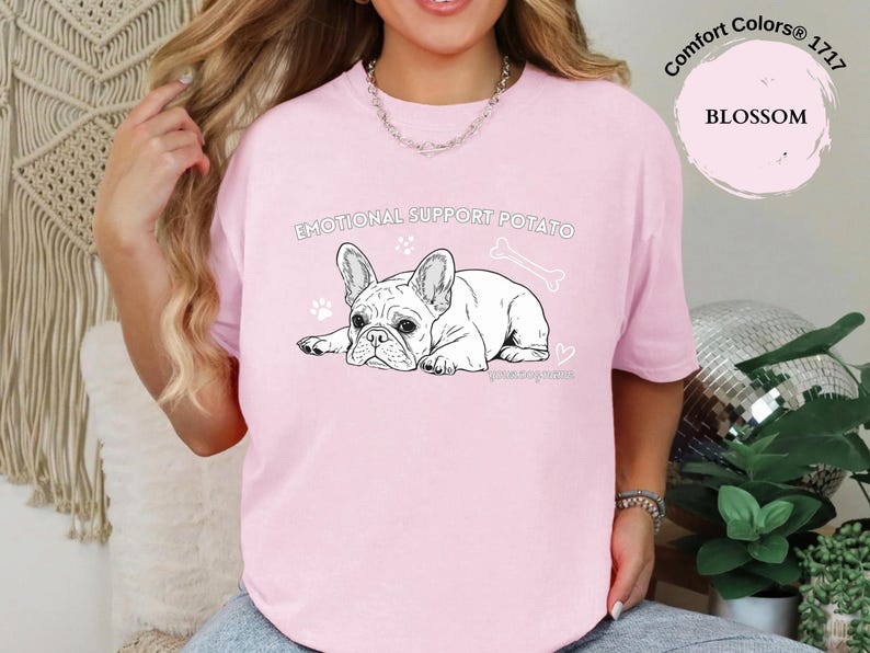 May include: A pale pink t-shirt featuring a black and white French Bulldog illustration with the text "EMOTIONAL SUPPORT POTATO". The shirt also has paw prints, a bone, and the word "BLOSSOM".