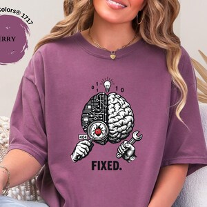 May include: A berry-colored t-shirt featuring a graphic of a brain split between a circuit board and organic tissue. The design includes a magnifying glass over a red bug, a lightbulb, binary code, a wrench, the number 404, and the text FIXED.