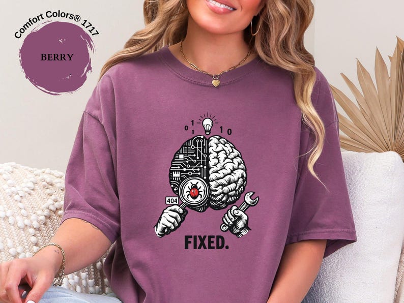 May include: A berry-colored t-shirt featuring a graphic of a brain split between a circuit board and organic tissue. The design includes a magnifying glass over a red bug, a lightbulb, binary code, a wrench, the number 404, and the text FIXED.