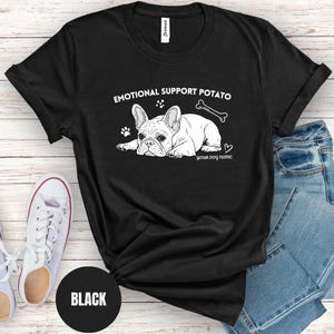 May include: Black t-shirt featuring a white illustration of a French Bulldog, a bone, and the text "EMOTIONAL SUPPORT POTATO" and "YOUR DOG NAME". The shirt is styled with blue jeans and white trainers.