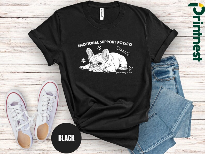 May include: Black t-shirt featuring a white illustration of a French Bulldog, a bone, and the text "EMOTIONAL SUPPORT POTATO" and "YOUR DOG NAME". The shirt is styled with blue jeans and white trainers.