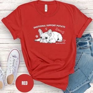May include: A red t-shirt featuring the text "EMOTIONAL SUPPORT POTATO" and a French Bulldog graphic. The design includes a bone, paw prints, and a heart. The words "your dog name" are below the dog. White trainers and blue jeans are in the background.