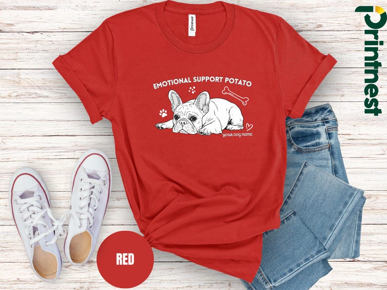 May include: A red t-shirt featuring the text "EMOTIONAL SUPPORT POTATO" and a French Bulldog graphic. The design includes a bone, paw prints, and a heart. The words "your dog name" are below the dog. White trainers and blue jeans are in the background.