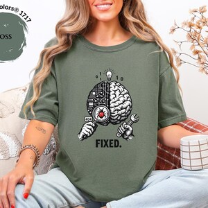 May include: A moss green t-shirt featuring a graphic of a brain split between a circuit board and organic tissue. One hand holds a magnifying glass over a red bug, while the other holds a wrench. Above is a lightbulb and binary code, with the text FIXED.