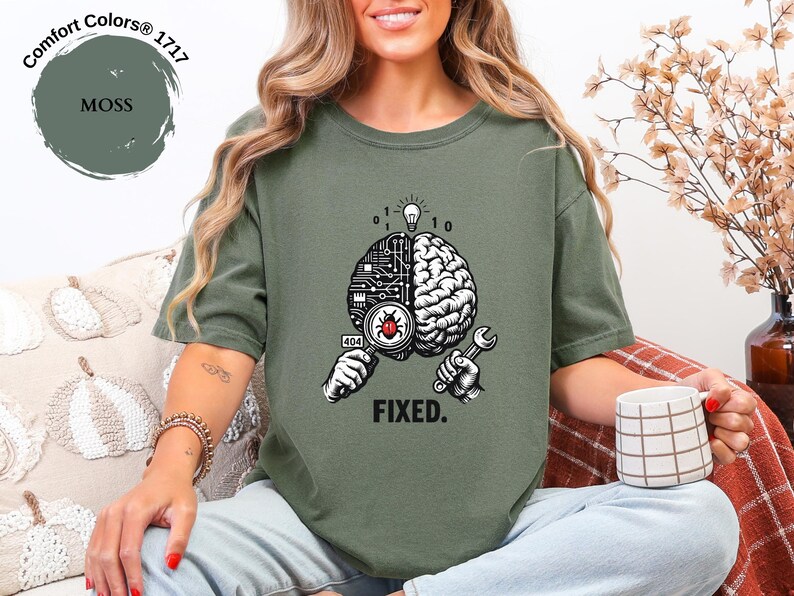 May include: A moss green t-shirt featuring a graphic of a brain split between a circuit board and organic tissue. One hand holds a magnifying glass over a red bug, while the other holds a wrench. Above is a lightbulb and binary code, with the text FIXED.