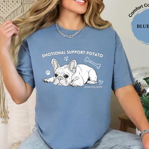 May include: Blue jean Comfort Colors t-shirt featuring a French Bulldog graphic and the text "EMOTIONAL SUPPORT POTATO". The design includes a bone, paw prints, and a heart. The text "your dog name" is below the dog.