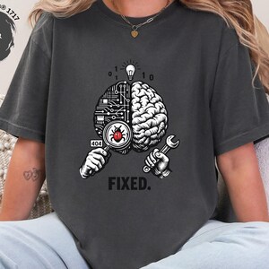 May include: A charcoal gray t-shirt featuring a graphic of a brain split into a circuit board and organic side. One hand holds a magnifying glass over a red bug with a 404 tag, while the other holds a wrench. A lightbulb and binary code appear above, with the word FIXED. printed below.