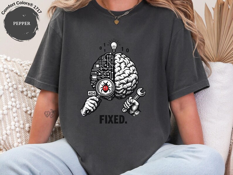 May include: A charcoal gray t-shirt featuring a graphic of a brain split into a circuit board and organic side. One hand holds a magnifying glass over a red bug with a 404 tag, while the other holds a wrench. A lightbulb and binary code appear above, with the word FIXED. printed below.