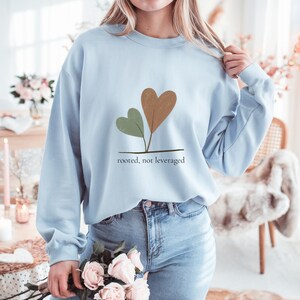 May include: A light blue sweatshirt featuring a graphic of two heart-shaped leaves in brown and green, with the text "rooted, not leveraged" below. The sweatshirt is long-sleeved and made of a soft material.