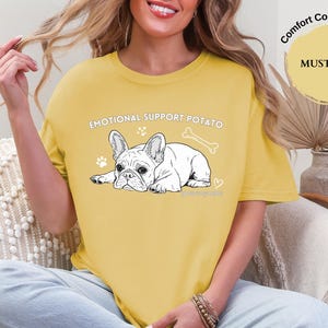 May include: Mustard yellow t-shirt featuring the text "EMOTIONAL SUPPORT POTATO" and a black and white illustration of a French Bulldog. The design includes a bone, paw prints, and a heart.