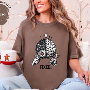 May include: An espresso brown short-sleeve t-shirt featuring a graphic of a brain split between a circuit board and organic tissue. One hand holds a magnifying glass over a red bug, while the other holds a wrench. Text reads 404 and FIXED.