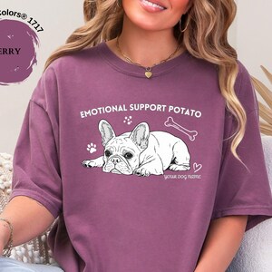 May include: Berry-coloured t-shirt featuring a French Bulldog graphic with the text "EMOTIONAL SUPPORT POTATO". The design includes a bone, paw prints, and a heart. The text "your dog name" is below the dog.