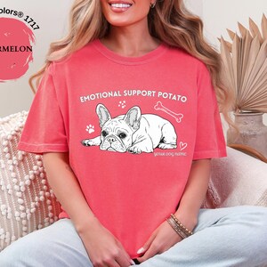 May include: A coral-coloured t-shirt with the text "EMOTIONAL SUPPORT POTATO" and a graphic of a white French Bulldog. The design includes a bone, paw prints, and a heart.