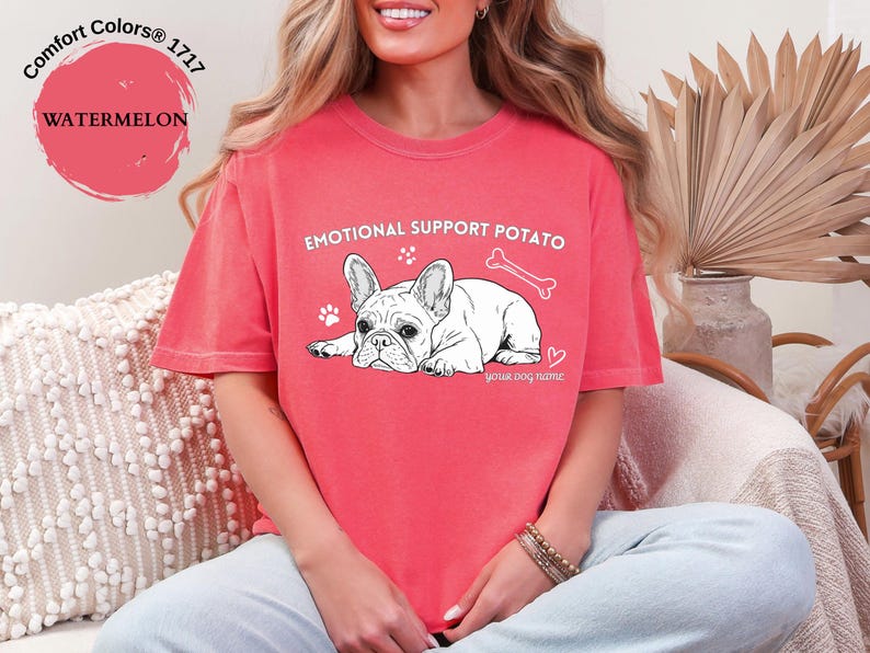 May include: A coral-coloured t-shirt with the text "EMOTIONAL SUPPORT POTATO" and a graphic of a white French Bulldog. The design includes a bone, paw prints, and a heart.