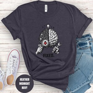 May include: A heather midnight navy t-shirt featuring a graphic of a brain split into a circuit board and organic side. One hand holds a magnifying glass over a red bug, while the other holds a wrench. Above is a lightbulb and the text FIXED.