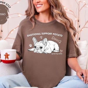 May include: Espresso-coloured t-shirt with the text "EMOTIONAL SUPPORT POTATO" and a graphic of a French Bulldog. The shirt also features a bone, paw prints, and a heart. A white mug with a gingerbread man is held in the hand.